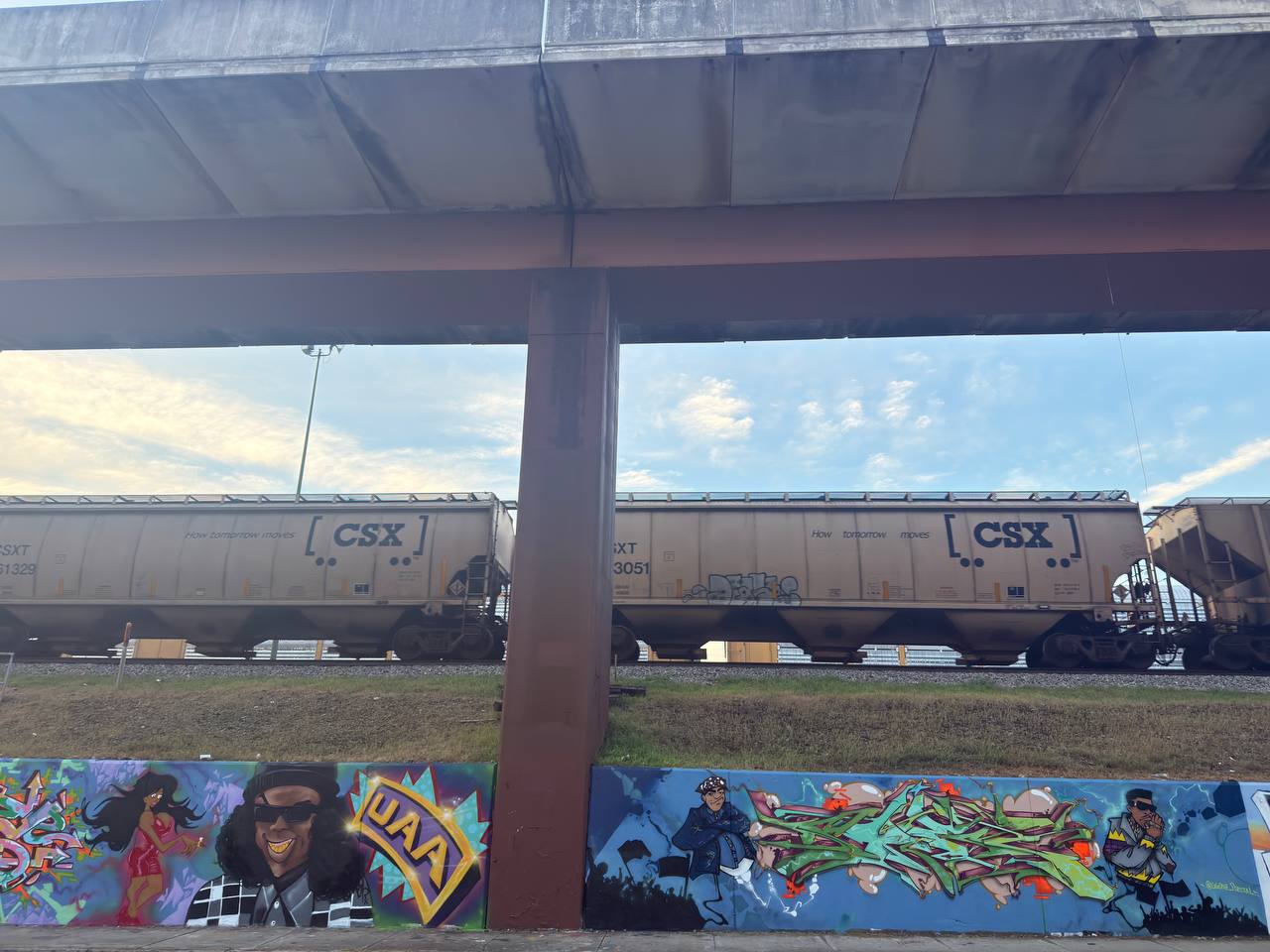 A freight train under an overpass beside a graffiti wall